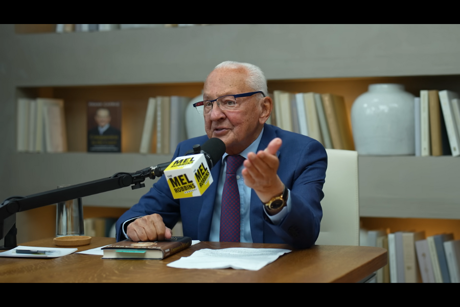 An older man in a blue suit speaks passionately into a microphone during a podcast recording. The mic has a "Mel Robbins Podcast" logo, and books line the shelves behind him.
