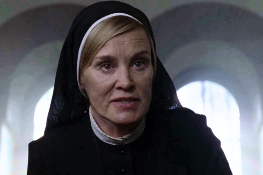 A stern-looking woman in a black nun’s habit and white collar sits in a dimly lit room with arched windows behind her.