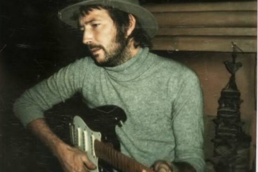 Eric Clapton playing a black electric guitar while wearing a grey turtleneck and wide-brimmed hat in a dimly lit room.