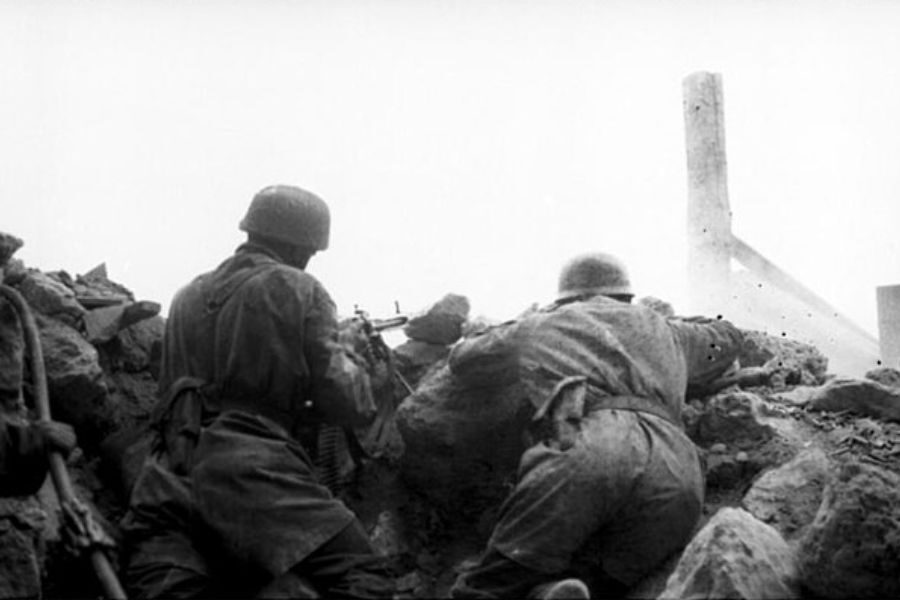 Two soldiers in combat gear crouch behind rubble with their rifles aimed forward amid the smoke and ruins of a battlefield.