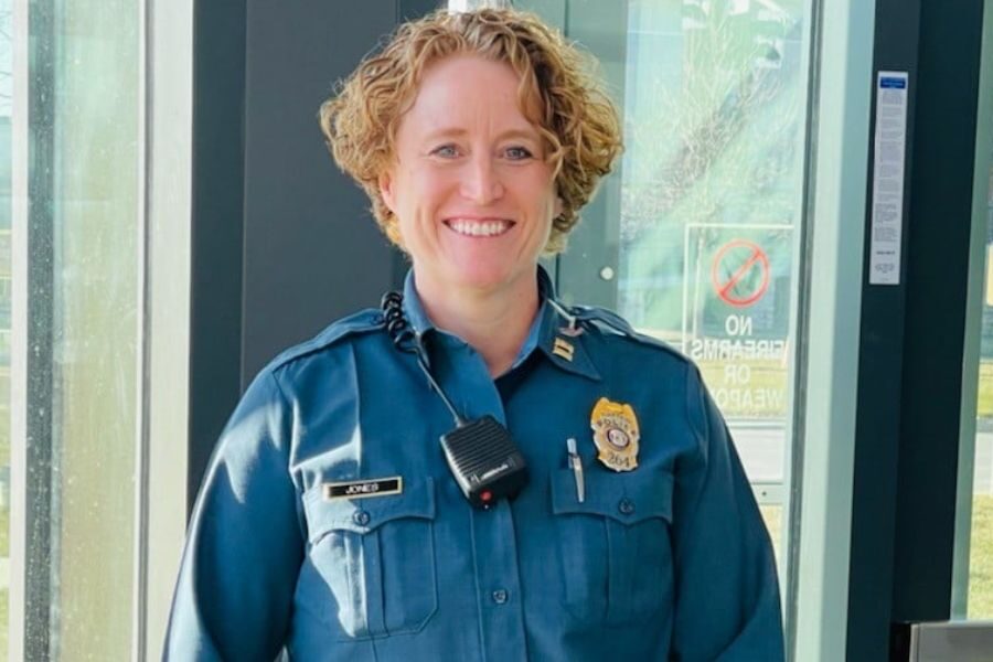 Smiling female police officer in uniform with a name tag reading "Jones" stands in front of glass doors.