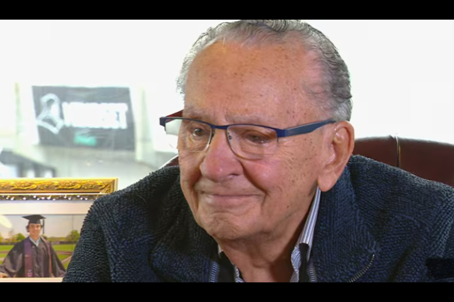Judge Frank Caprio smiles gently while sitting at a desk with a framed graduation photo nearby, suggesting a sentimental or reflective moment.
