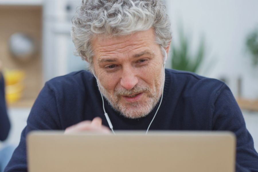 An older man with gray hair and earbuds is speaking during a video call, looking at his laptop screen with a slight smile.