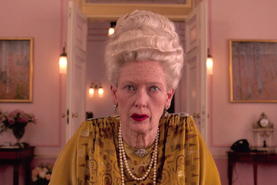 An elderly woman with dramatic white hair and deep red lipstick sits in an opulently decorated pink room, wearing gold brocade and layered pearls.