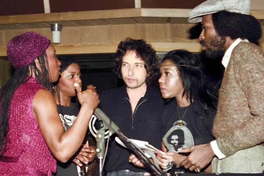 Bob Dylan in a recording studio surrounded by vocalists and musicians, all engaged in conversation or singing near microphones.