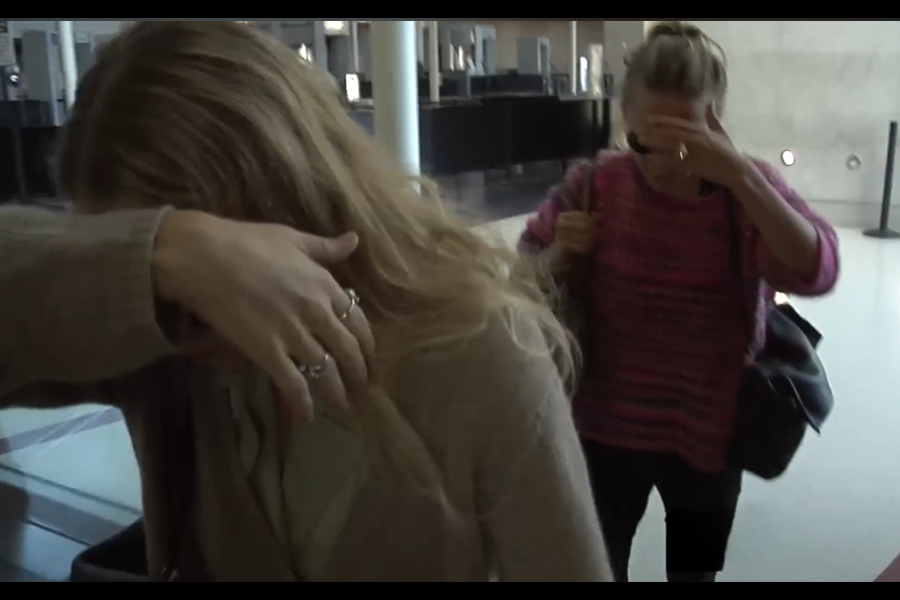 Two women walk indoors with their heads down, covering their faces with their hands to avoid cameras or attention.