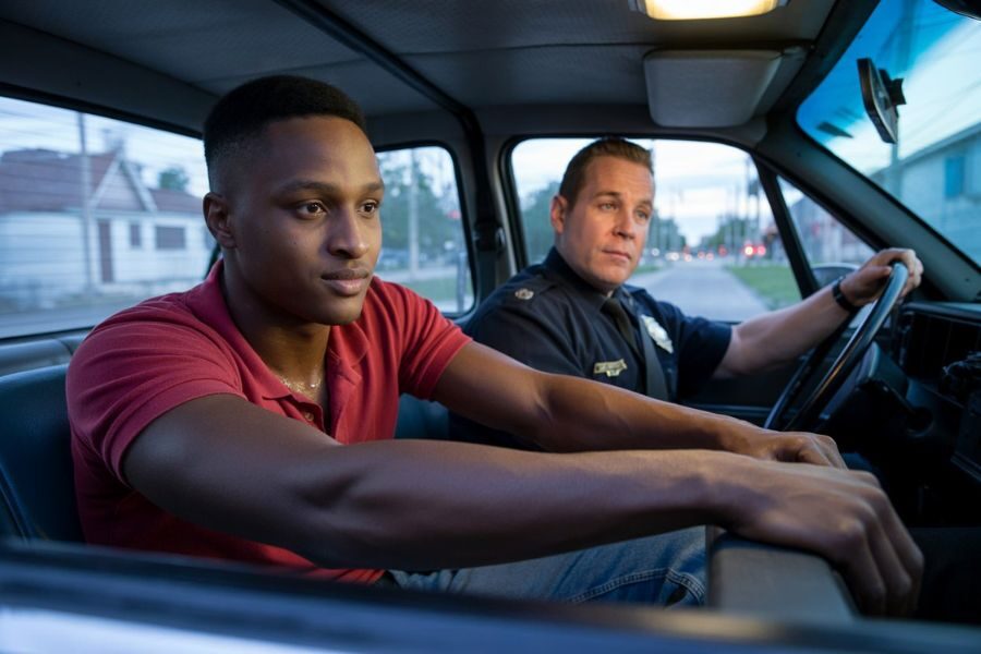 A young man and a police officer sit together inside a car, both looking focused and calm, possibly during a ride-along or conversation.