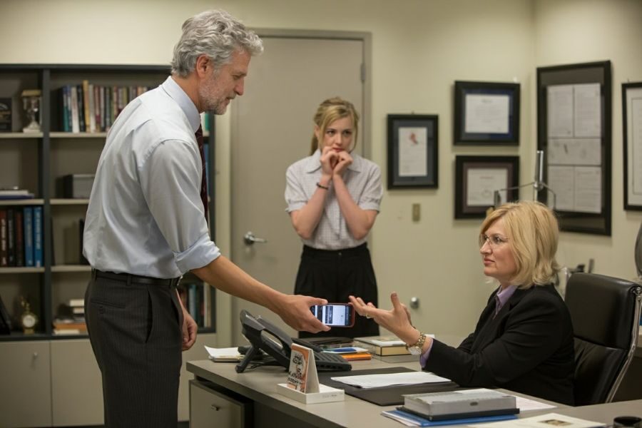 A man hands his phone to a serious-looking woman behind a desk, while a concerned young woman stands in the background.