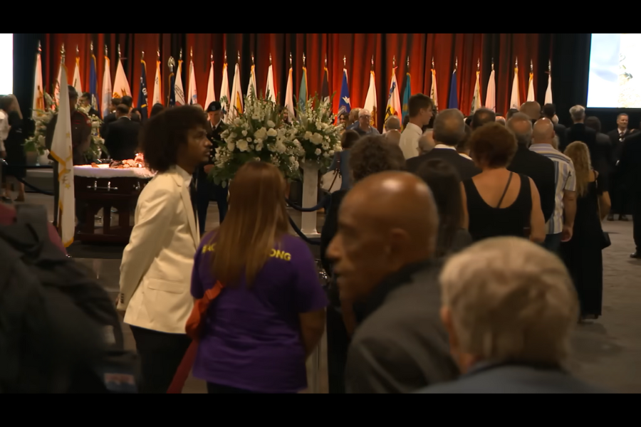 A large crowd attends a memorial service in a hall adorned with white floral arrangements and many flags; a casket is visible at the front.