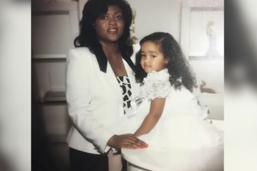 A woman in a white suit stands next to a young girl in a white frilly dress, posing together inside a home.