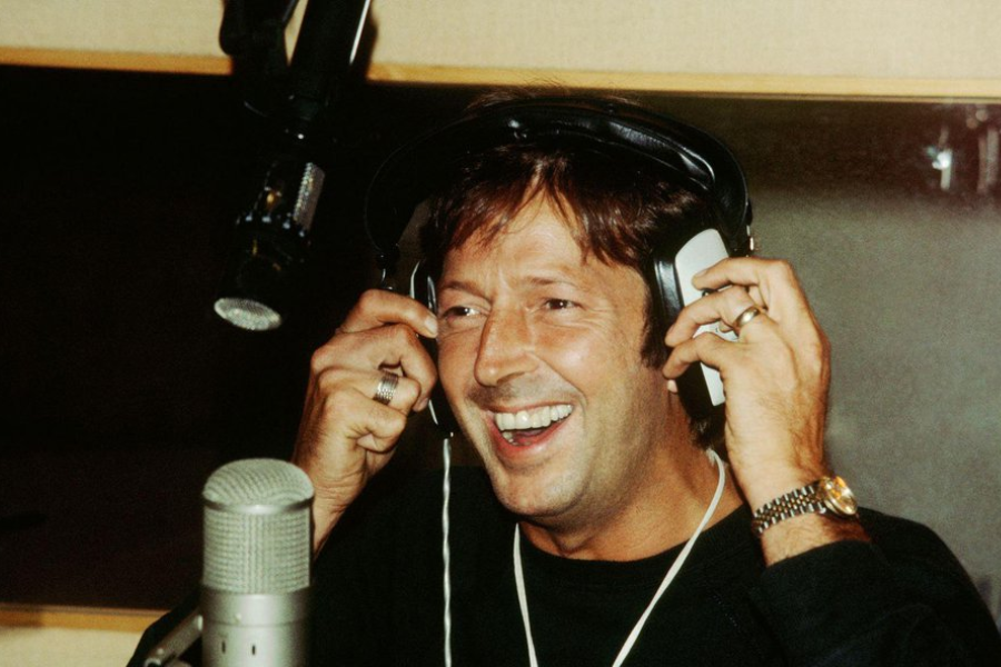Eric Clapton laughing while recording vocals in a studio, wearing large headphones and rings on both hands.