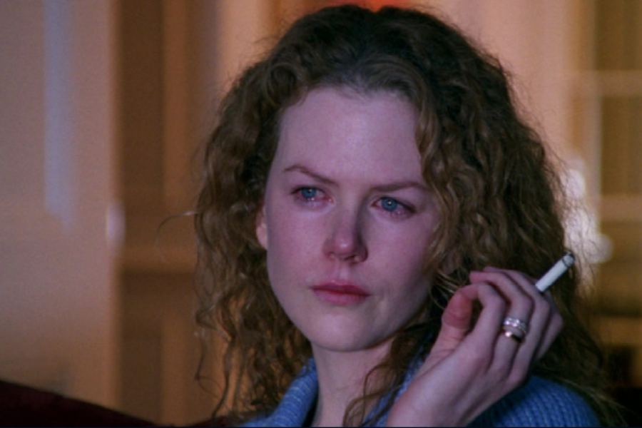 A woman with red-rimmed eyes and curly hair holds a cigarette and stares solemnly into the distance while wearing a blue robe.