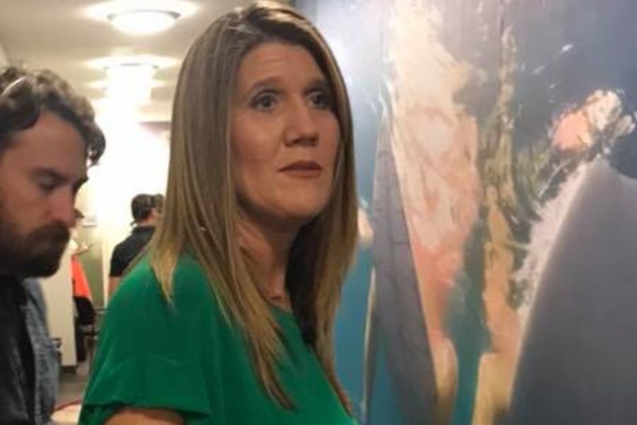 A woman in a green blouse stands indoors with a soft, concerned expression as a man stands slightly behind her.