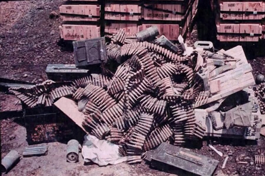Large tangled heap of ammunition belts, crates, and various munitions piled outdoors beside stacked wooden boxes.