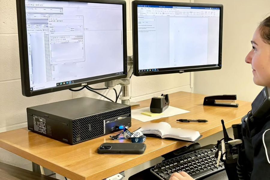 Female officer seated at a desk working on a desktop computer with two monitors displaying forms and documents.