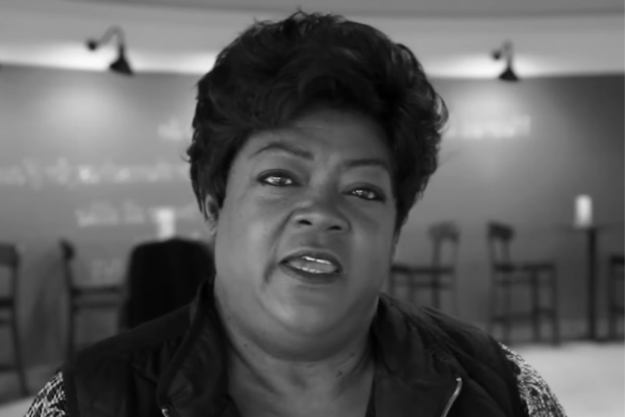 A black and white image of a woman speaking, seated in a room with high-top tables in the background.