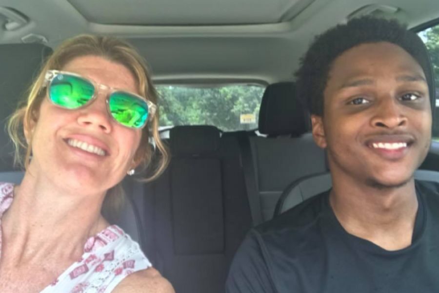 A smiling woman in sunglasses takes a selfie with a young man in the passenger seat, both appearing happy inside a parked car.