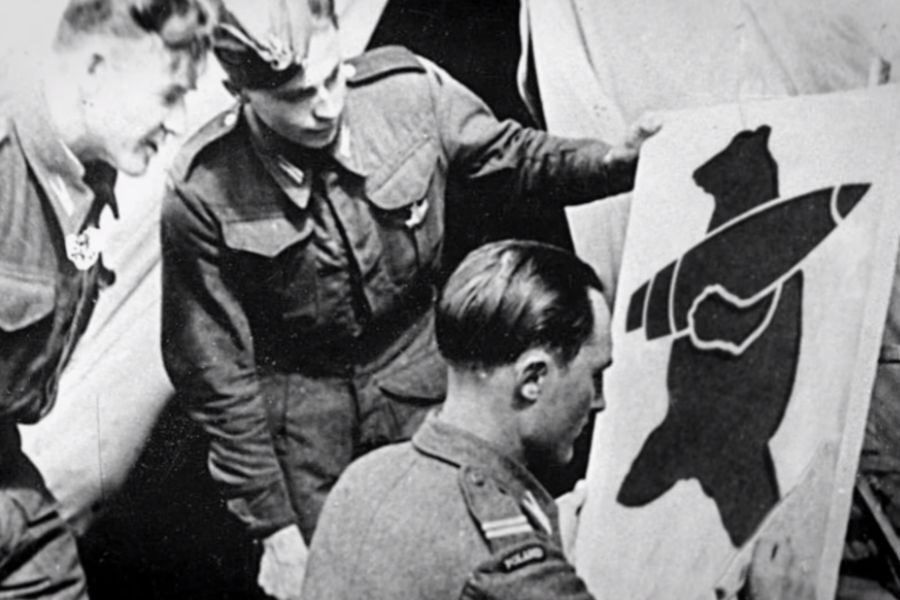 Three uniformed soldiers gather around a painted banner showing a black bear carrying an artillery shell across its shoulder.