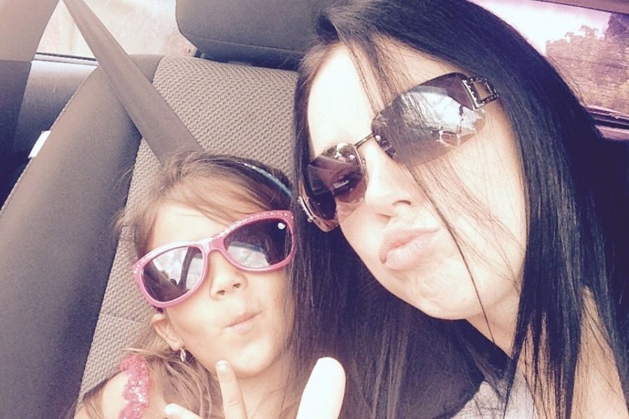 Woman and young girl pose together in a car, both wearing sunglasses and making playful faces.