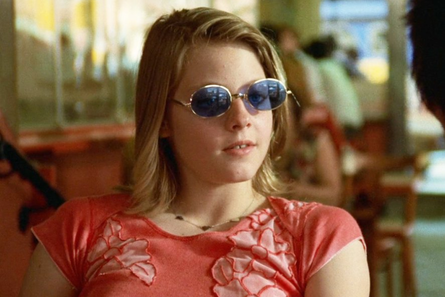 A young woman in round blue sunglasses and a coral shirt with floral embroidery lounges in a diner, speaking casually.