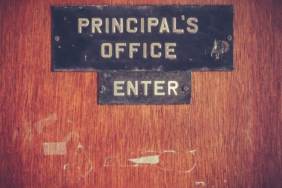 A weathered wooden door with a sign that reads “Principal’s Office Enter,” showing signs of age and wear.