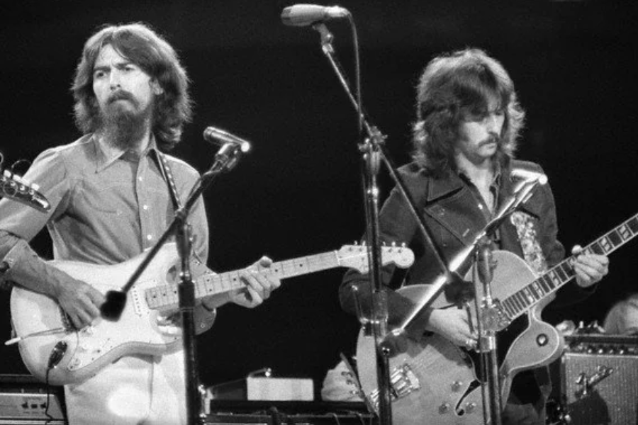 George Harrison and Eric Clapton play electric guitars side-by-side during a live performance, surrounded by microphones.