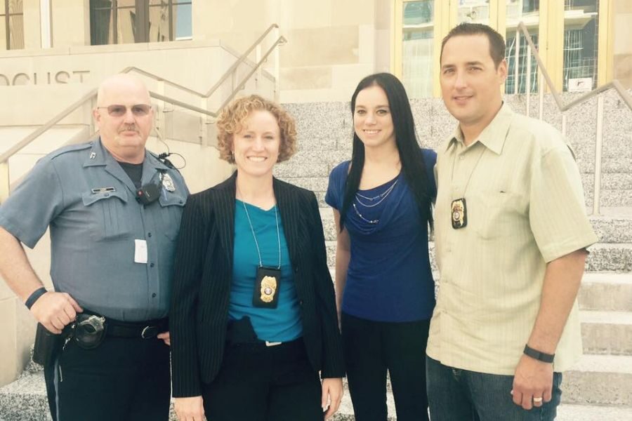 Group of four law enforcement officers and personnel stand together on courthouse steps posing for a photo.