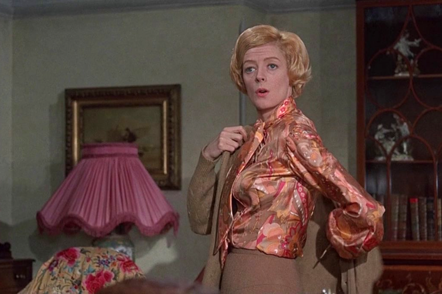 A woman with styled blonde hair dramatically pulls on a printed blouse, standing in a traditionally decorated room with books and floral accents.