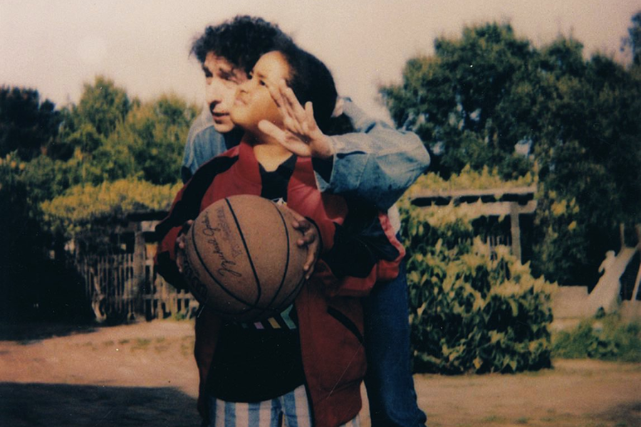 A man, possibly Bob Dylan, helps a young girl aim while holding a basketball in an outdoor setting with trees and fencing in the background.