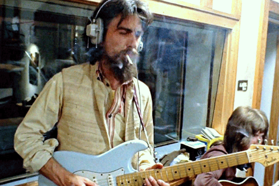 George Harrison smokes a cigarette while playing a white electric guitar in a recording studio, headphones over his long hair.