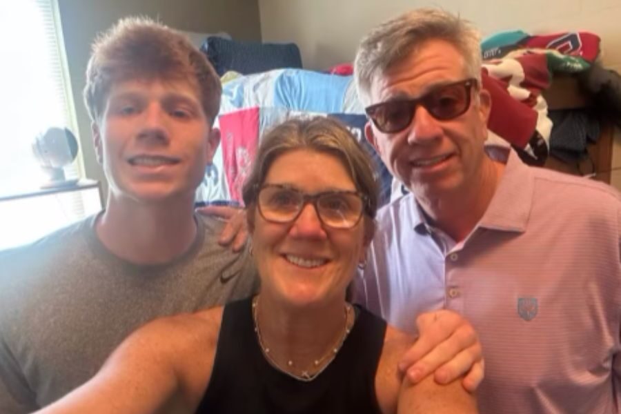 A smiling woman stands between a teenage boy and a man in sunglasses, posing for a selfie in a bedroom with colorful bedding in the background.