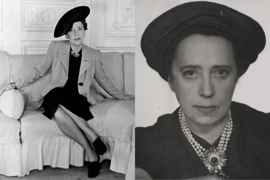 Woman in a structured blazer and dark dress sits on a couch wearing a tilted wide-brimmed hat and brooch, with legs crossed. Close-up shows her serious expression and elegant pearl necklace.