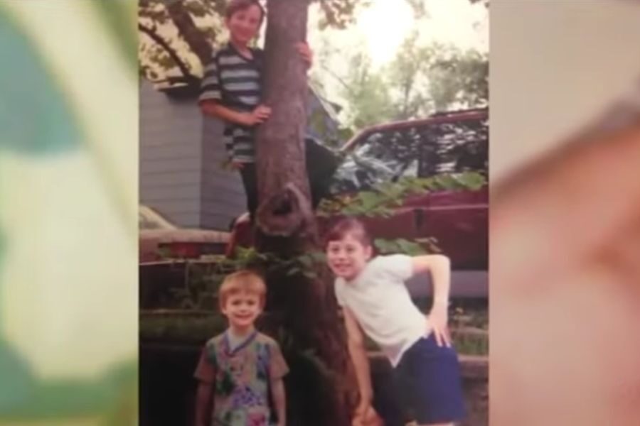 Three children pose outdoors by a tree, with one boy climbing it and the others smiling below.