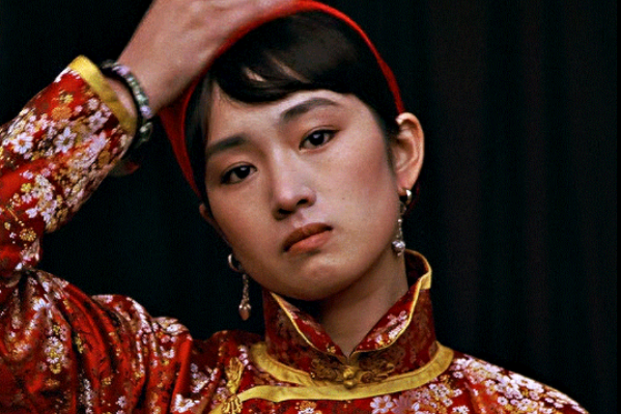 A young woman in a red and gold embroidered outfit looks pensively forward while adjusting her hair, with dangling earrings and minimal expression.
