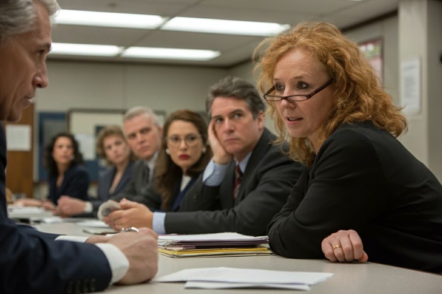 A woman in a black suit speaks during a tense school board meeting, surrounded by serious and attentive colleagues.