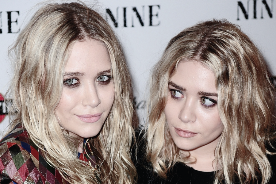Two young twin girls with blonde hair and bangs smile brightly in matching shiny outfits at what appears to be a red carpet event.