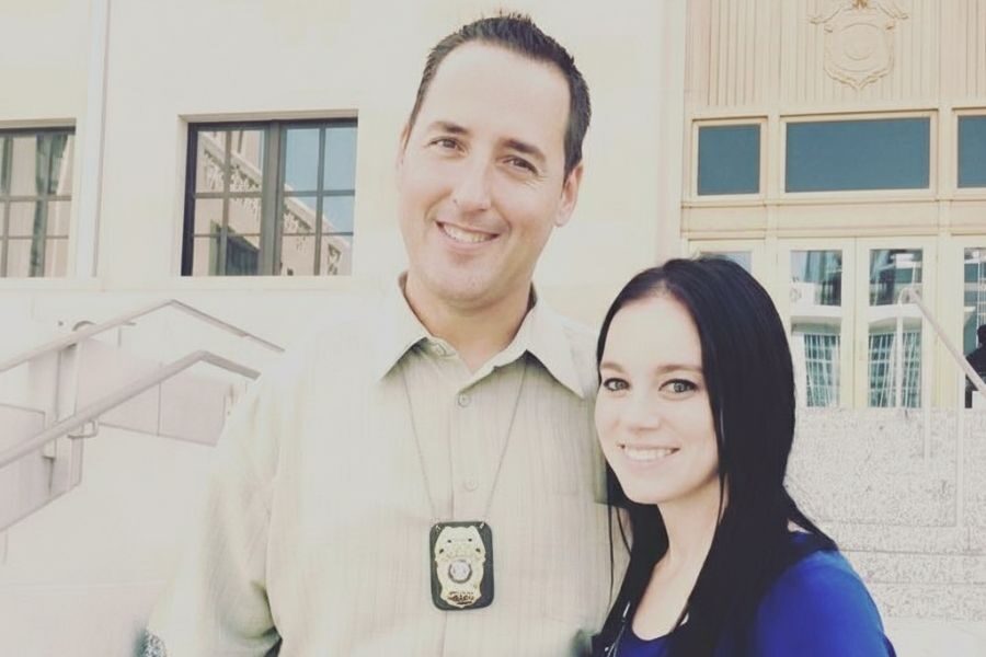 Smiling man wearing a badge around his neck poses with a woman in front of a government building.