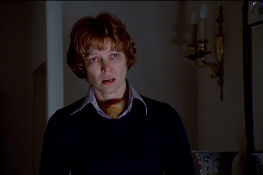 A disheveled woman with a fearful expression stands in a dim hallway, wearing a navy sweater and collared shirt with a scarf tucked in.