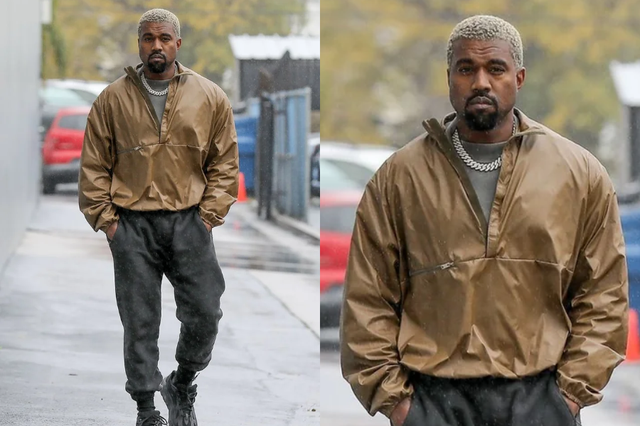 Man with platinum blonde hair walks down a wet street in a loose tan windbreaker, dark joggers, and chunky black boots, wearing a thick silver chain. Close-up highlights his serious expression.