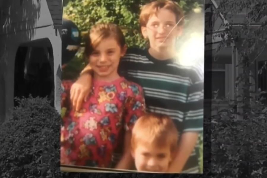 Three smiling children pose outdoors, with one girl wearing a colorful cartoon shirt and the boys standing behind her.