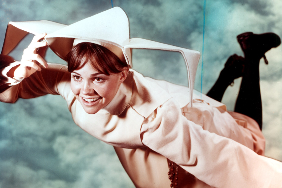 A smiling young woman dressed as a nun in a cream-colored flying habit appears suspended in midair against a cloudy sky, playfully holding her headpiece.