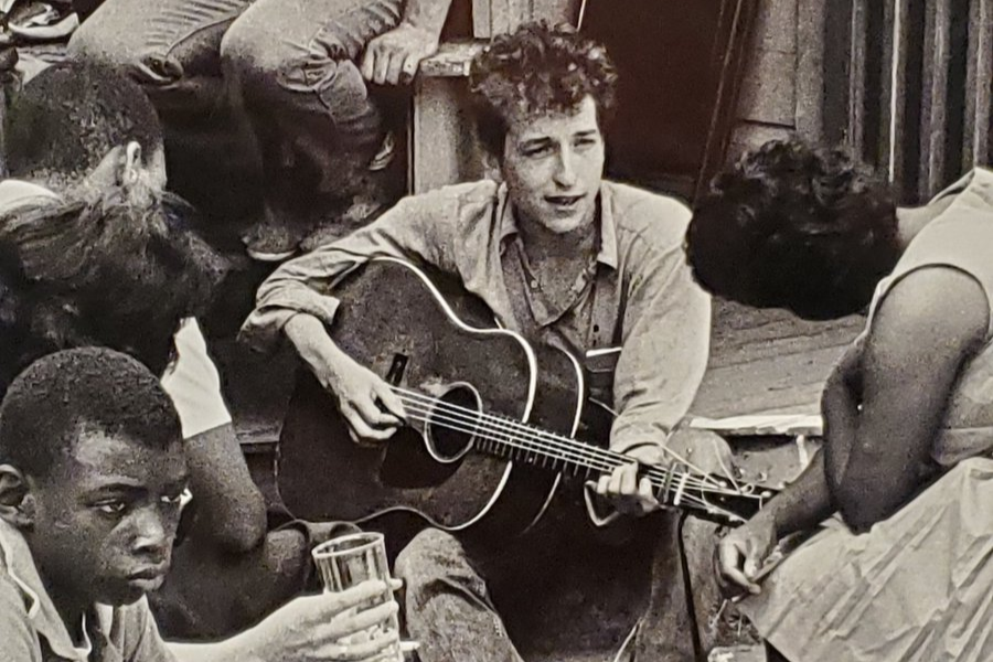  Alt text: A young man playing an acoustic guitar among a group of seated people, engaging with them in what looks like an informal outdoor performance or gathering.