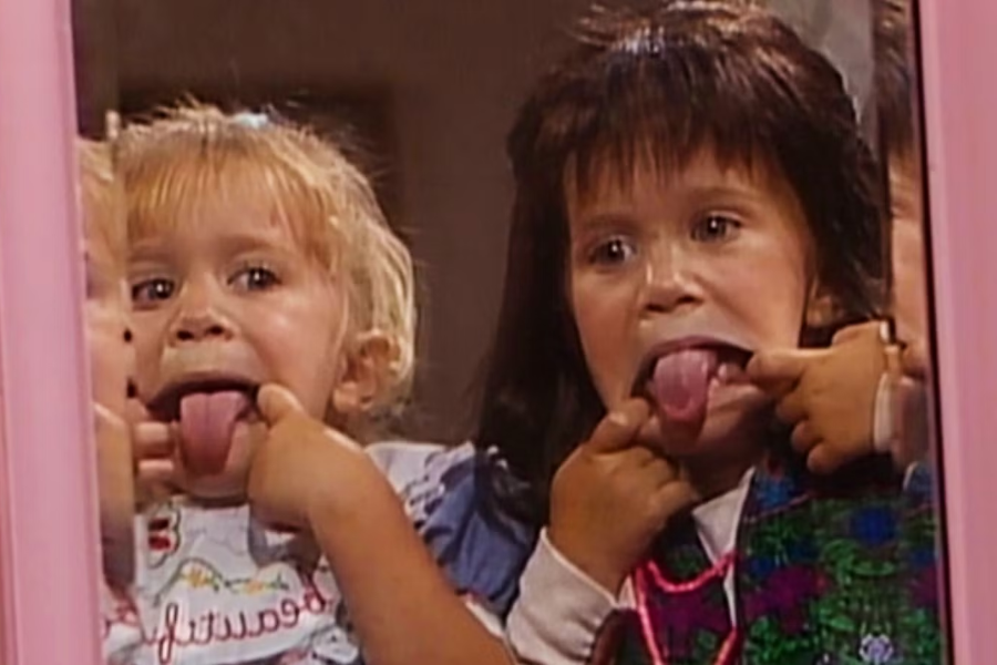 A young blonde girl sticks her tongue out and pulls a funny face in front of a mirror next to a dark-haired girl mimicking the same gesture.