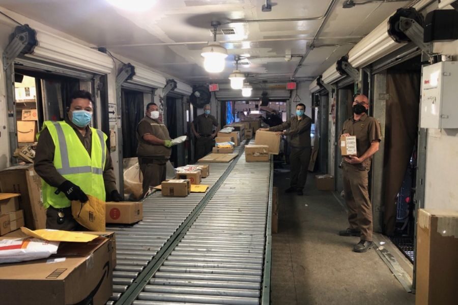 Several masked delivery workers in brown uniforms sorting packages on a conveyor belt inside a large delivery truck.