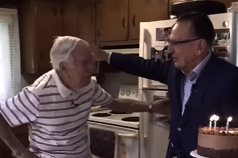 An elderly man in a striped shirt greets another man holding a birthday cake with lit candles, sharing a warm moment in a cozy kitchen.
