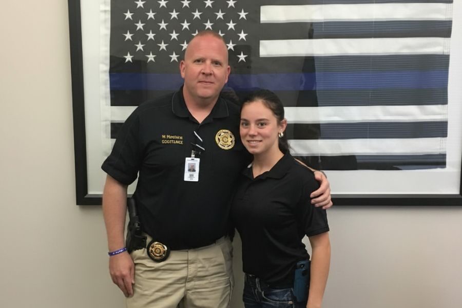 Uniformed officer with arm around a young woman stands in front of a black and white American flag with a blue line.