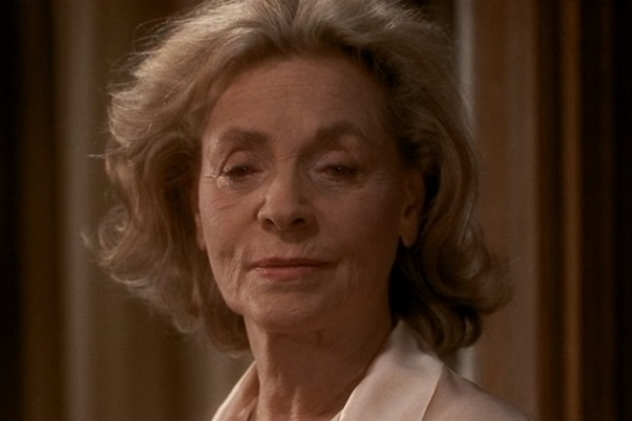 An older woman with soft waves of silver hair gives a subtle, knowing smile while wearing a white blouse in a warmly lit room.