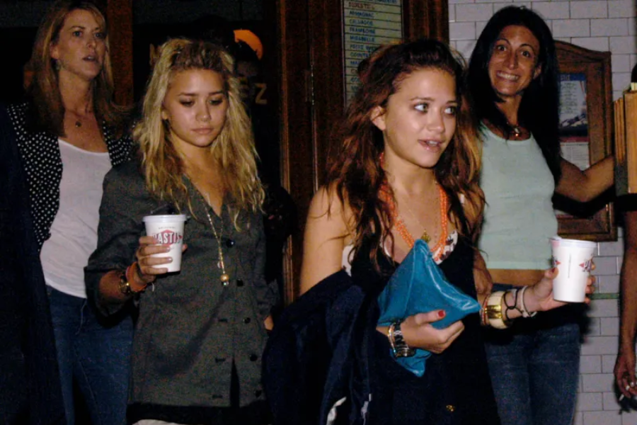 Mary-Kate and Ashley Olsen are seen leaving a venue at night with drinks in hand, surrounded by others. They look tired and casual in dim lighting.