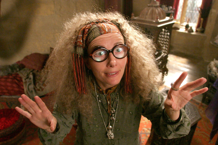 A wide-eyed woman with frizzy hair and oversized glasses raises her hands mid-gesture, wearing layers of jewelry and a headscarf in a cluttered, mystical room.