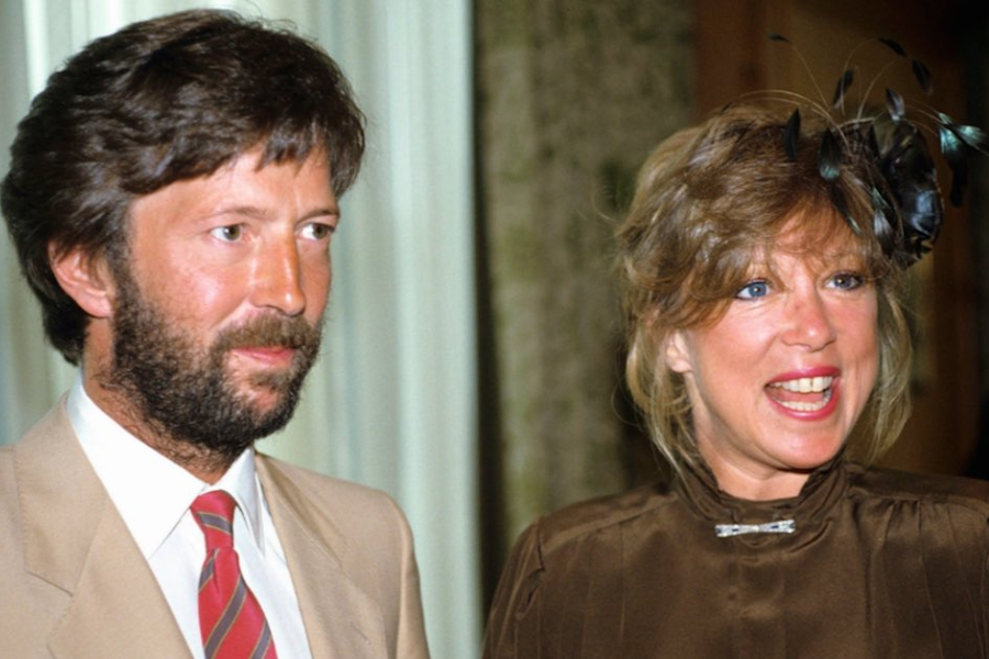 Eric Clapton and Pattie Boyd dressed formally at an event, with Clapton in a tan suit and red-striped tie and Boyd smiling in a brown dress with a feathered hairpiece.